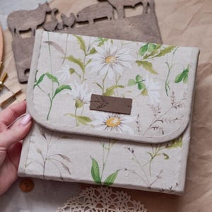 May include: A square fabric pouch featuring a beige background with a botanical print of white daisies and green foliage. The pouch has a fold-over flap secured with a rectangular brown leather-like label centered on the front with a small bird icon.