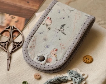 May include: A fabric needle case with a light blue bird and botanical print, featuring a grey polka-dot border and a dark button closure. The case is accompanied by metal scissors, small wooden buttons, and two skeins of blue embroidery thread.
