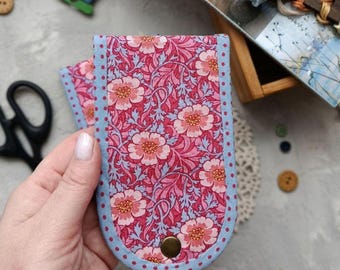 May include: A rectangular fabric case with rounded bottom edges, featuring a pink and red floral pattern on a light blue background. The case is trimmed with light blue fabric with small red dots and secured with a single brass-colored metal snap.