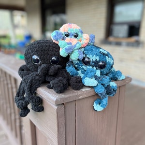 May include: Three crocheted octopus plushies in different colors. The octopus on the left is black, the middle octopus is pink and blue, and the octopus on the right is blue and green.