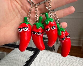 Puede incluir: Cuatro llaveros de pimiento rojo con tallos verdes y caras de dibujos animados. Cada llavero tiene una cadena y un anillo plateados. Los pimientos tienen ojos blancos, pupilas negras y mejillas rosadas. Los llaveros se sostienen frente a un fondo blanco.