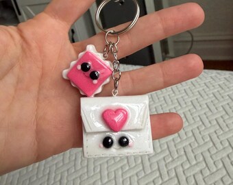 Puede incluir: Un llavero hecho a mano con dos colgantes: un cuadrado rosa con una cara sonriente y un sobre blanco con un corazón rosa. Los colgantes tienen ojos negros y están unidos a un llavero plateado.