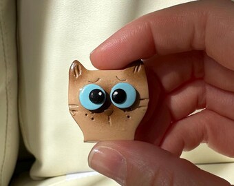 Puede incluir: Pequeña figura en forma de gato marrón con grandes ojos azules. La figura tiene un diseño sencillo con detalles visibles como las orejas, la nariz y los bigotes. El gato se sostiene entre los dedos, mostrando su tamaño y forma.