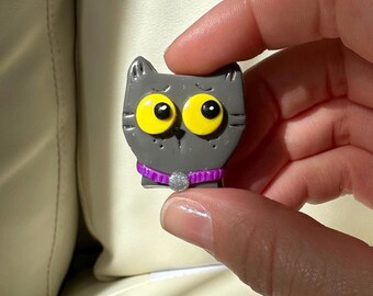 Puede incluir: Un dije gris con forma de gato con grandes ojos amarillos y un collar morado con un acento plateado. El dije se sostiene en una mano, mostrando su pequeño tamaño y diseño detallado. El gato tiene una apariencia linda y de dibujos animados.