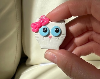 Puede incluir: Una figura blanca con forma de gato con un lazo rosa, ojos azules y una nariz rosa. El gato tiene pupilas negras y pequeños puntos blancos en la cara. La figura se sostiene en una mano.