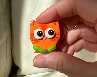 Puede incluir: Una pequeña figura en forma de gato naranja con grandes ojos negros y blancos y un collar verde. El gato tiene una expresión ligeramente triste y un pequeño adorno redondo en su collar. La figura se sostiene en una mano.