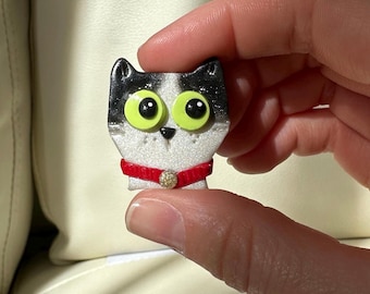 Puede incluir: Pequeña figura con forma de gato con cabeza negra y blanca, ojos verdes grandes y collar rojo. El collar tiene un adorno dorado. La figura está sujeta por los dedos de una persona.