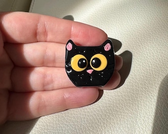 Puede incluir: Un objeto decorativo negro con forma de gato con grandes ojos amarillos, una nariz rosa y orejas internas rosas. El gato tiene un acabado brillante y un diseño sencillo y de dibujos animados. El artículo es probablemente un alfiler, un imán o un elemento decorativo.