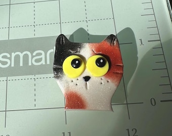 Puede incluir: Un objeto decorativo con forma de gato, blanco, negro y naranja. El gato tiene grandes ojos amarillos con pupilas negras. El objeto está sobre una superficie verde con medidas en centímetros.