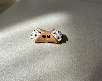 Puede incluir: Un cannoli de dibujos animados con una cara sonriente. La cáscara de la pastelería es de color marrón claro, rellena de crema blanca y chispas de chocolate. El cannoli en miniatura es un objeto decorativo.