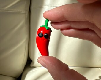 Puede incluir: Una pequeña figura de pimiento rojo con un tallo verde y ojos negros. El pimiento tiene una cara sonriente y se sostiene entre dos dedos. La figura es probablemente un objeto decorativo o un juguete pequeño.