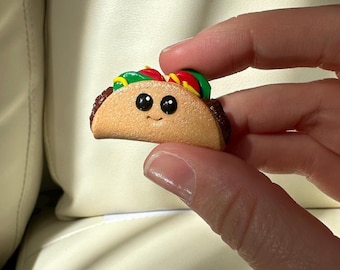 Puede incluir: Un dije de taco sonriente en miniatura con una concha marrón claro y bordes marrón oscuro. El taco está cubierto con ingredientes verdes, rojos y amarillos. El dije tiene ojos grandes y negros.