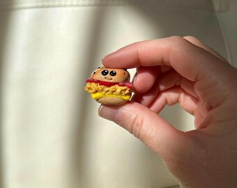 Puede incluir: Una hamburguesa en miniatura hecha a mano con una cara sonriente. La hamburguesa presenta un panecillo amarillo, una hamburguesa frita, tomate rojo y una rebanada de carne rosa. La hamburguesa se sostiene entre los dedos sobre un fondo neutro.