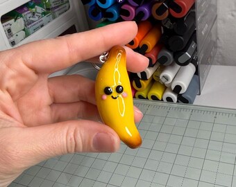 Puede incluir: Un llavero amarillo con forma de plátano con una cara sonriente y mejillas rosadas. El llavero tiene un acabado brillante y un pequeño anillo de metal para sujetarlo a llaves o a un bolso. El fondo incluye bolígrafos y rotuladores de colores.