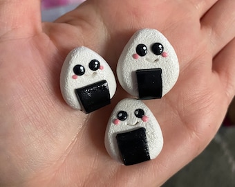 Puede incluir: Tres onigiri, una bola de arroz japonesa, blancas y negras, con caras de dibujos animados lindas. Los onigiri tienen ojos negros, mejillas rosadas y una tira de alga nori negra.