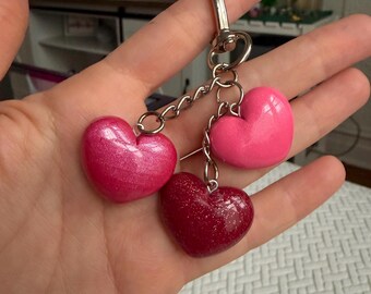 Puede incluir: Un llavero con tres dijes en forma de corazón en tonos de rosa y rojo. Los corazones están unidos a una cadena y un cierre plateados. Un corazón tiene purpurina, los otros son de color sólido.