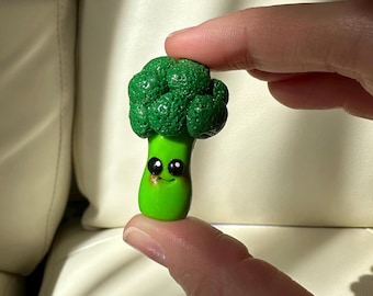 Puede incluir: Una caprichosa figura de brócoli pintada a mano. La cabeza del brócoli es de un verde oscuro texturizado, mientras que el tallo es de un verde brillante. La figura tiene una cara sonriente con ojos negros, añadiendo un toque juguetón a esta pieza única.