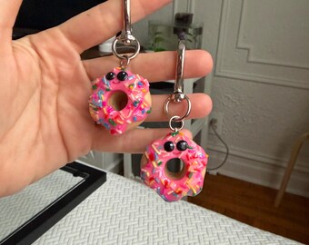Puede incluir: Dos llaveros en forma de rosquilla rosa con chispas de colores y ojos negros. Cada llavero tiene un cierre de color plateado. Los donuts son sostenidos por una mano.