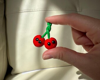 Puede incluir: Un par de amuletos rojos con forma de cereza con ojos negros y tallos verdes. Las cerezas se sostienen entre los dedos sobre un fondo neutro. Los amuletos tienen un acabado brillante y un diseño caprichoso.