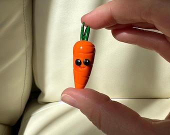 Puede incluir: Una pequeña figura de zanahoria naranja con una cara sonriente y hojas verdes. La zanahoria tiene ojos negros y se sostiene entre dos dedos. La figura es un artículo artesanal caprichoso.