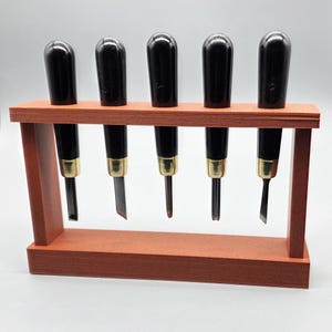 May include: Five wood carving tools with black handles and gold ferrules, presented in a reddish-brown wooden rack. The blades are visible, showing various cutting profiles. These tools are designed for woodworking and carving projects.