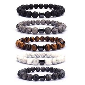 Op de afbeelding: Vijf kralenarmbanden met een zwarte hematietkraal in het midden van elke armband. De armbanden zijn gemaakt van zwarte lavasteen, grijs marmer, tijgeroog, witte howliet en grijs agaat.
