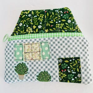 Peut inclure: Une pochette matelassée en forme de maison avec un toit floral vert et un corps à carreaux gris et blanc. La pochette présente des détails appliqués tels que des fenêtres, une porte et des plantes en pot. Une fermeture éclair et une bordure vertes complètent le design.