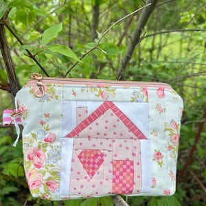 May include: A rectangular fabric pouch featuring a quilted house design in shades of pink and white. The pouch is decorated with a floral pattern of pink roses and blue flowers on a white background, finished with a metallic zipper closure.