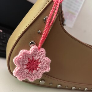 Op de afbeelding: Een lichtbruine handtas met zilveren studs en een roze gehaakte bloemhanger. De bloem heeft een donkerroze centrum en is met roze garen aan de tas bevestigd. De tas is gemaakt van een glad materiaal.