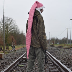 Op de afbeelding: Een persoon met een lange, roze gehaakte muts met witte rand, staand op een spoorweg. De muts steekt ver boven het hoofd uit. De persoon draagt een jas met patroon en een groene broek.
