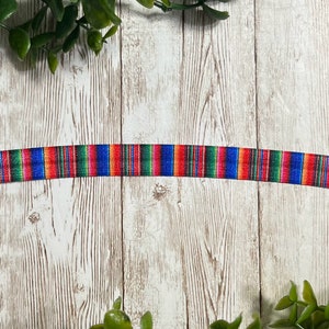 May include: A colorful ribbon with a striped pattern in red, blue, green, and yellow. The ribbon is displayed against a white wooden background with green foliage in the corners. The ribbon is likely used for crafting or decorative purposes.