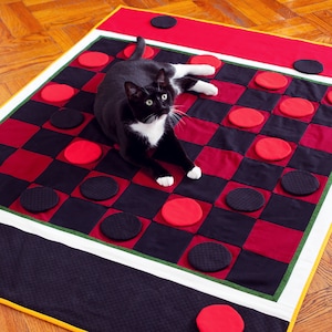 Puede incluir: Un gato blanco y negro sentado en una colcha de estilo tablero de ajedrez rojo y negro. La colcha tiene círculos negros y rojos cosidos en ella.