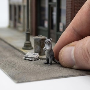 Pode incluir: Uma miniatura de guaxinim cinzento e preto em pé numa calçada cinzenta ao lado de um pequeno caixote do lixo de metal e uma pilha de jornais, posicionada em frente a um edifício modelo detalhado com um candeeiro de rua.