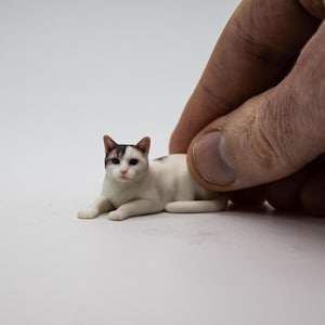 May include: A miniature figurine of a cat in a lying position. The cat has a white coat with dark gray patches on its head and ears, pointed ears, and dark eyes. The small sculpture is rendered with realistic details, capturing the feline's posture.