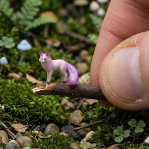 May include: A tiny pink and white fox ornament perched on a twig. The fox has a bushy tail and is set against a backdrop of green moss, small mushrooms, and other natural elements. The figurine is held by a person's finger.