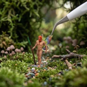 May include: A miniature figurine of a male fairy with orange hair, a red beard, and rainbow wings, holding a green-tipped spear. The fairy stands in a mossy forest setting with tiny mushrooms and a pair of tweezers.