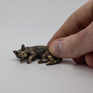 May include: A miniature figurine of a sleeping cat with a tortoiseshell coat featuring mottled black, brown, and orange fur patterns. The small, detailed sculpture depicts the feline curled up in a resting position with its head tucked down.