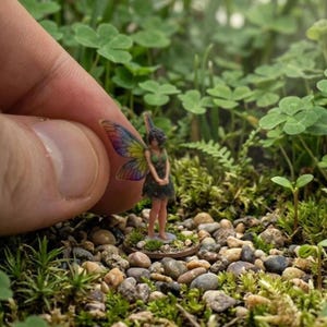 Op de afbeelding: Een miniatuur feeënbeeldje met kleurrijke vleugels en een groene jurk. De fee staat op een kleine, ronde basis te midden van een natuurlijke omgeving van kiezels, mos en klavers. De vleugels tonen een regenboog aan kleuren.