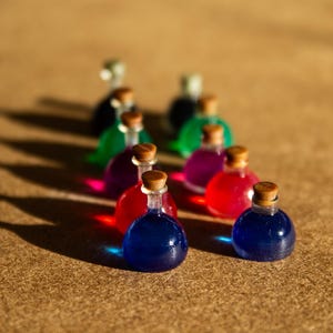 May include: A collection of miniature glass potion bottles with cork stoppers. The bottles contain colorful liquids in shades of blue, red, pink, green, and purple. The bottles are arranged on a brown surface, with shadows cast across the surface.