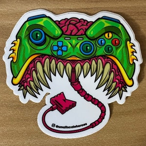 May include: A green and yellow monster-like video game controller with a pink cord and plug. The controller has a brain inside and a set of sharp teeth. The text "@smallscalheroes" is at the bottom of the image.