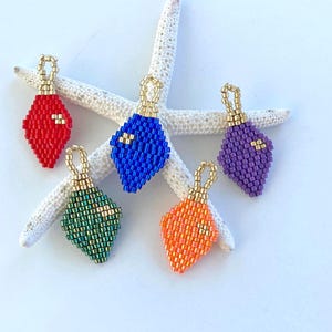May include: Five colorful beaded diamond-shaped ornaments with gold-colored loops. The ornaments are red, blue, green, orange, and purple. They are arranged on a white starfish against a white background.