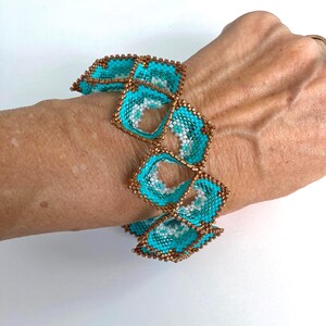 May include: A beaded bracelet featuring a series of turquoise and white square-shaped elements with a bronze-colored border. The bracelet is worn on a wrist, showcasing its intricate design and color contrast. The bracelet's design is geometric and eye-catching.