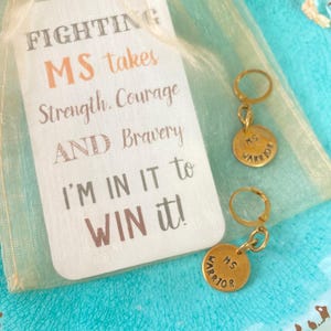 May include: A pair of gold-colored hoop earrings with round charms that read "MS WARRIOR". The earrings are displayed on a turquoise plate next to a tag with the text "FIGHTING MS takes Strength, Courage AND Bravery I'M IN IT to WIN it!"