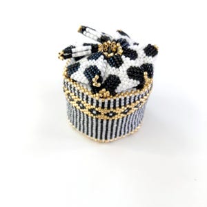 May include: A small, round beaded box with a lid. The lid is white with black spots, and the body is black and white striped. Gold-colored beads accent the box. The box is on a white background.