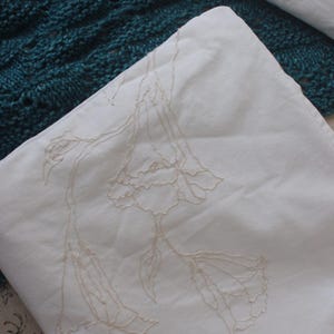 May include: A white pillowcase with embroidered floral design. The pillowcase is folded, revealing the intricate beige stitching of the flowers and leaves. A teal knitted blanket is visible in the background.