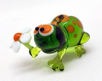Puede incluir: Figurita de vidrio de una tortuga verde con manchas naranjas, sosteniendo una margarita blanca con centro naranja. La tortuga tiene ojos grandes y expresivos, posando como si ofreciera la flor.