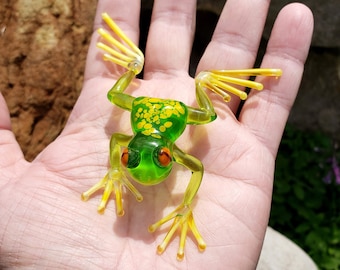 May include: A green glass frog figurine with yellow accents and orange eyes. The frog is sitting on a person's hand.