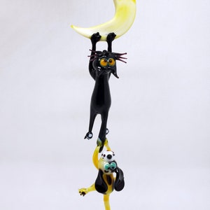 May include: A whimsical glass figurine featuring a black cat hanging from a yellow crescent moon. Below, a yellow and black dog figure dangles from the cat's paws. The figurine is suspended by a clear string, creating a playful, decorative piece.