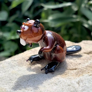 May include: A small glass figurine of a brown beaver with black eyes and a white tooth. The beaver is sitting on a light brown rock.