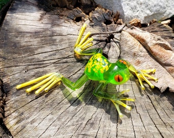 May include: A green glass frog with yellow accents and red eyes. The frog is sitting on a tree stump.
